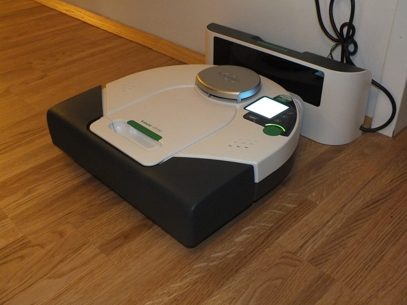 A robot vacuum parked on its charging base beside a wall on a wooden floor.