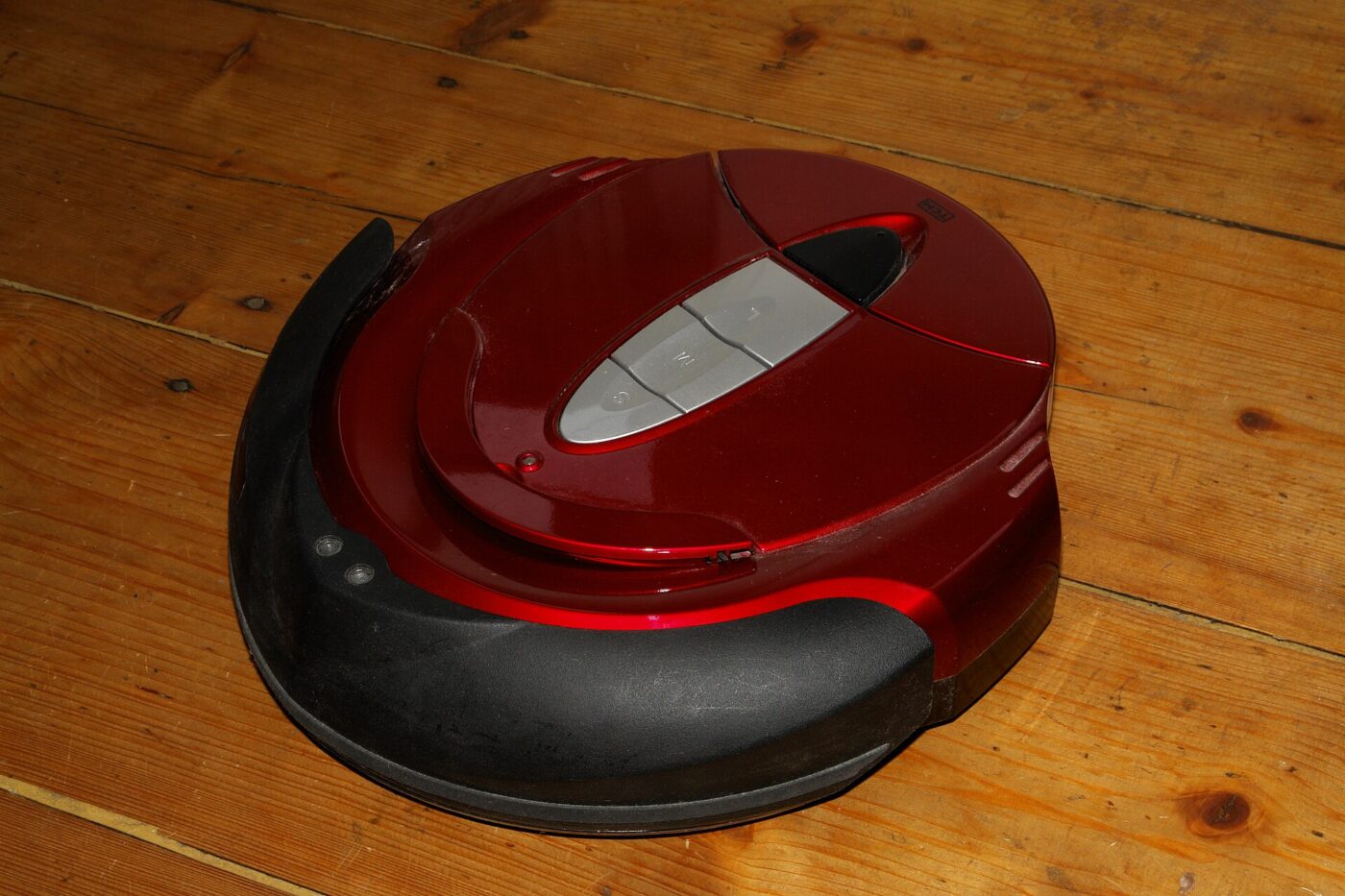 A round robot vacuum cleaner with a red top cover standing on a wooden floor.