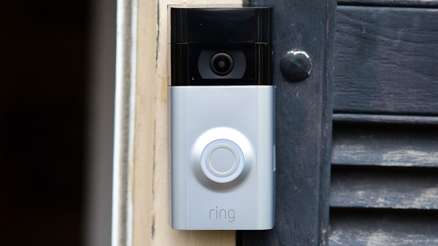 A video doorbell with a camera above its button, mounted on a door frame.