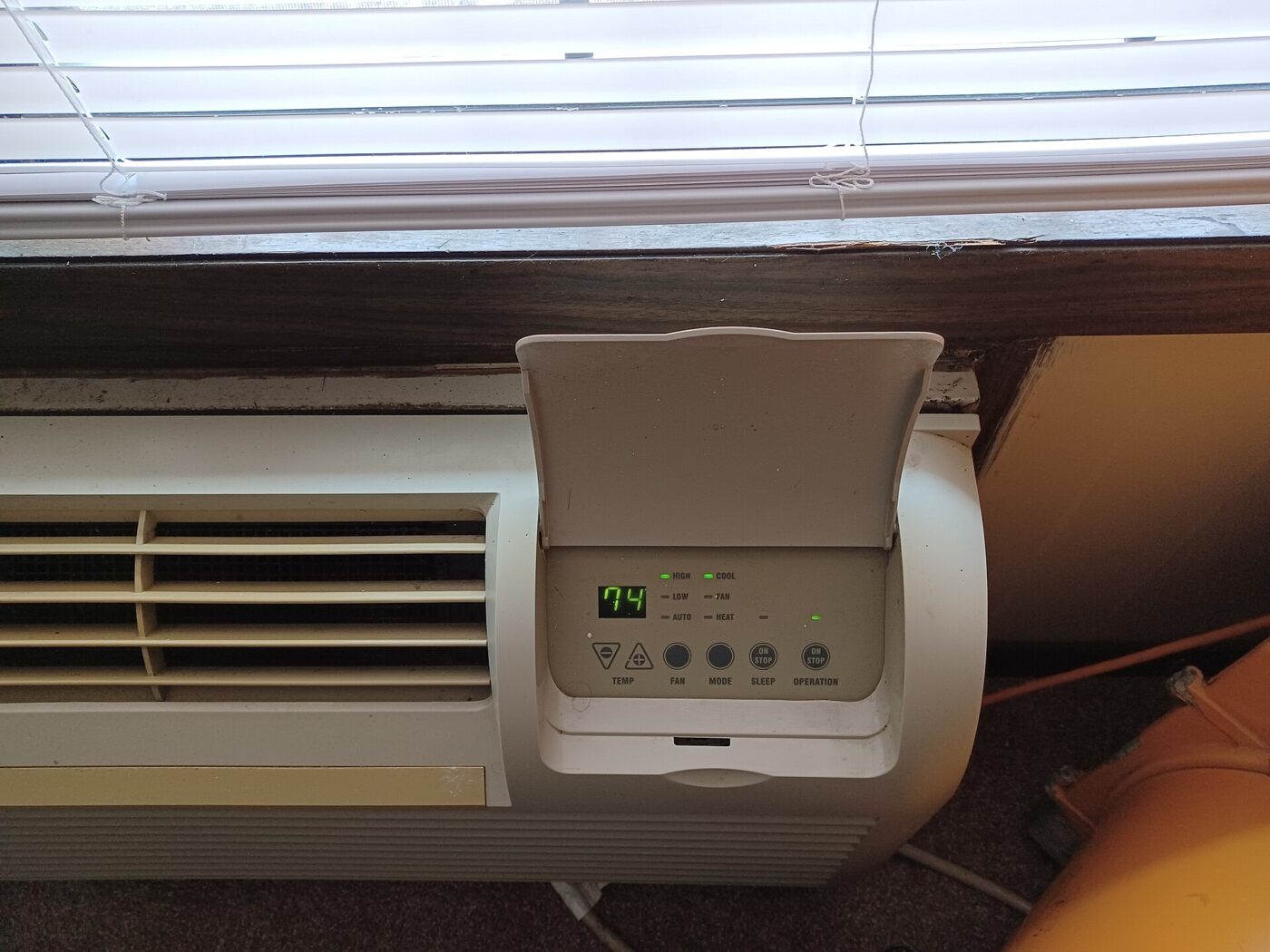 A through-the-wall room unit fitted under a window, with its control panel cover raised